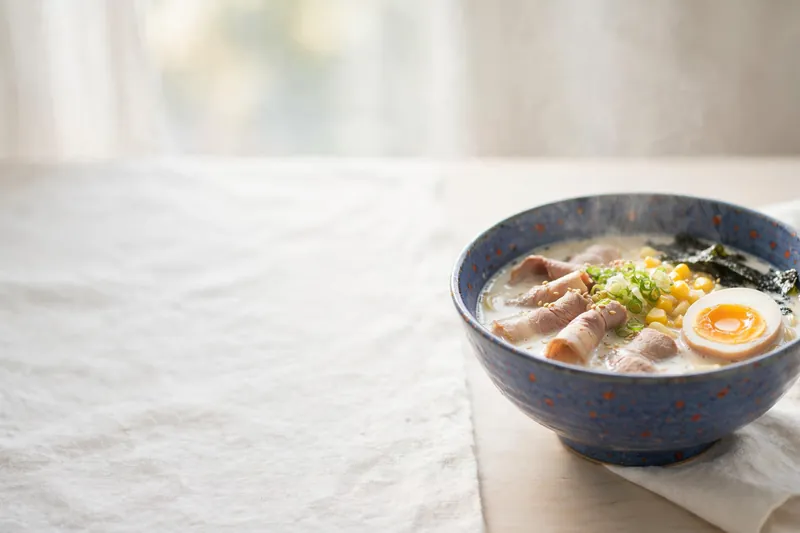 Illustration of a bowl of tonkotsu ramen on a pale wooden counter in morning light